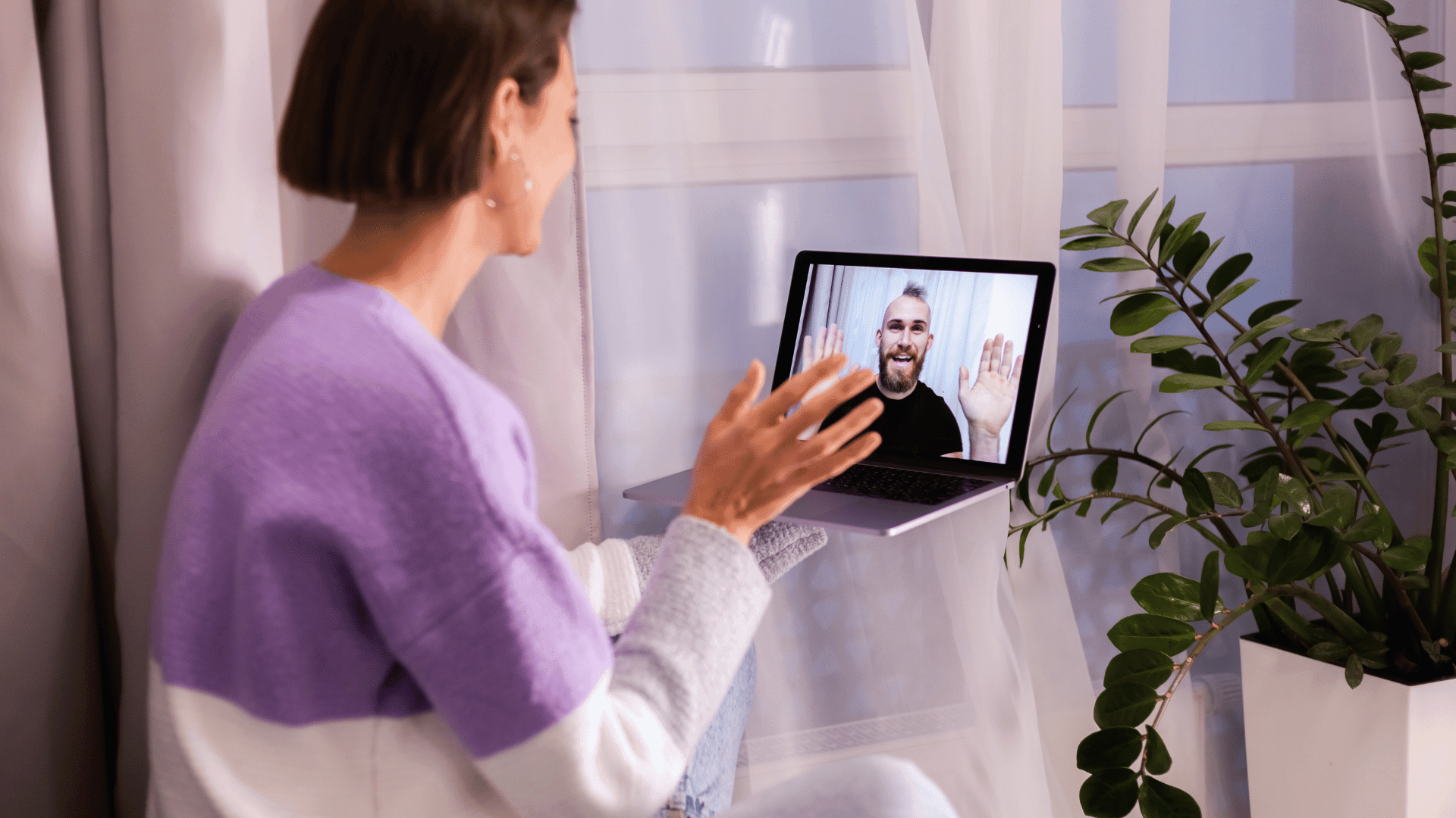 Woman in purple sweater waving and engaging with her therapist during an online DBT therapy session, with a plant visible in the background