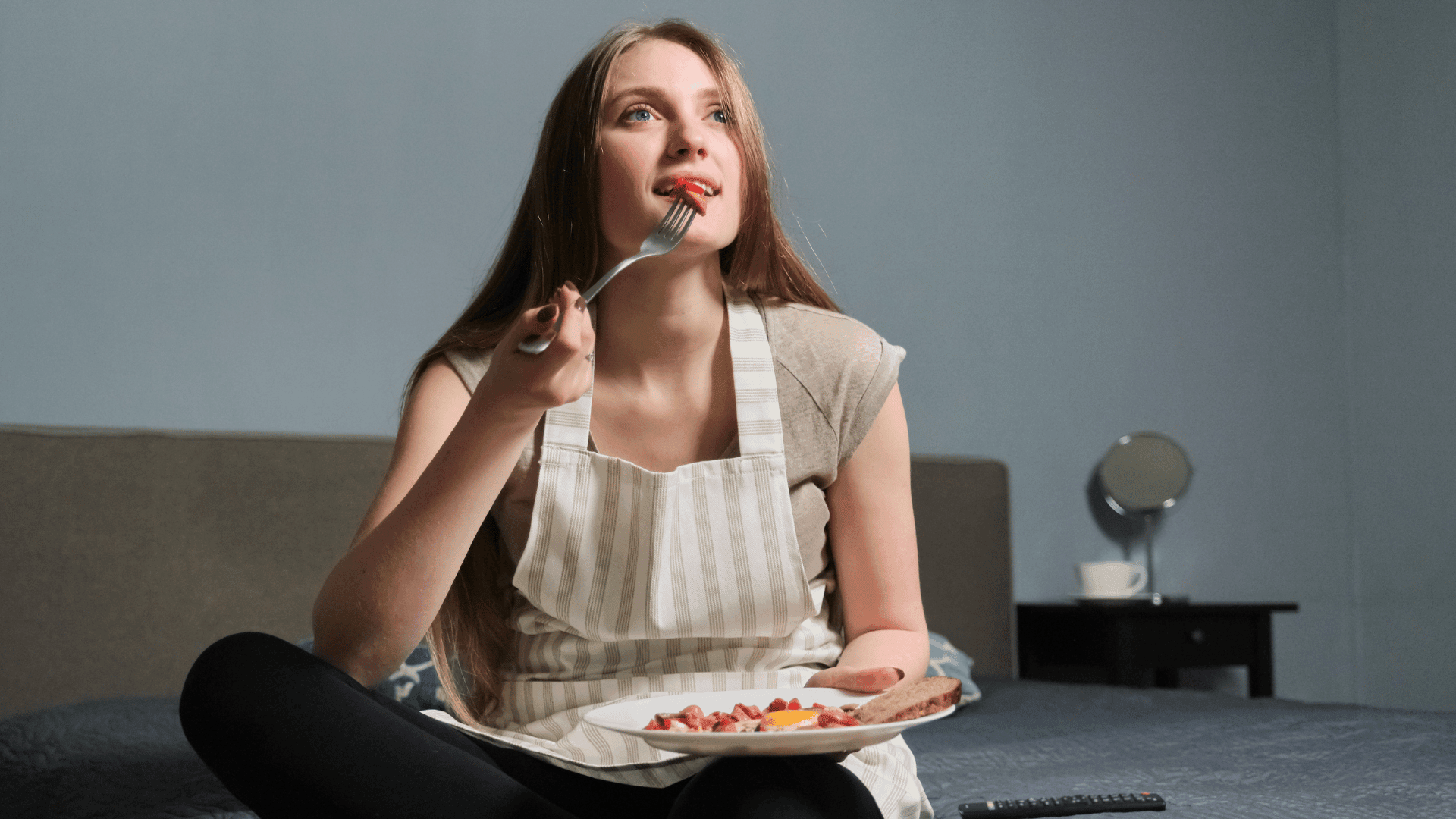 Young woman in striped apron enjoying a meal on her couch, demonstrating a positive relationship with food