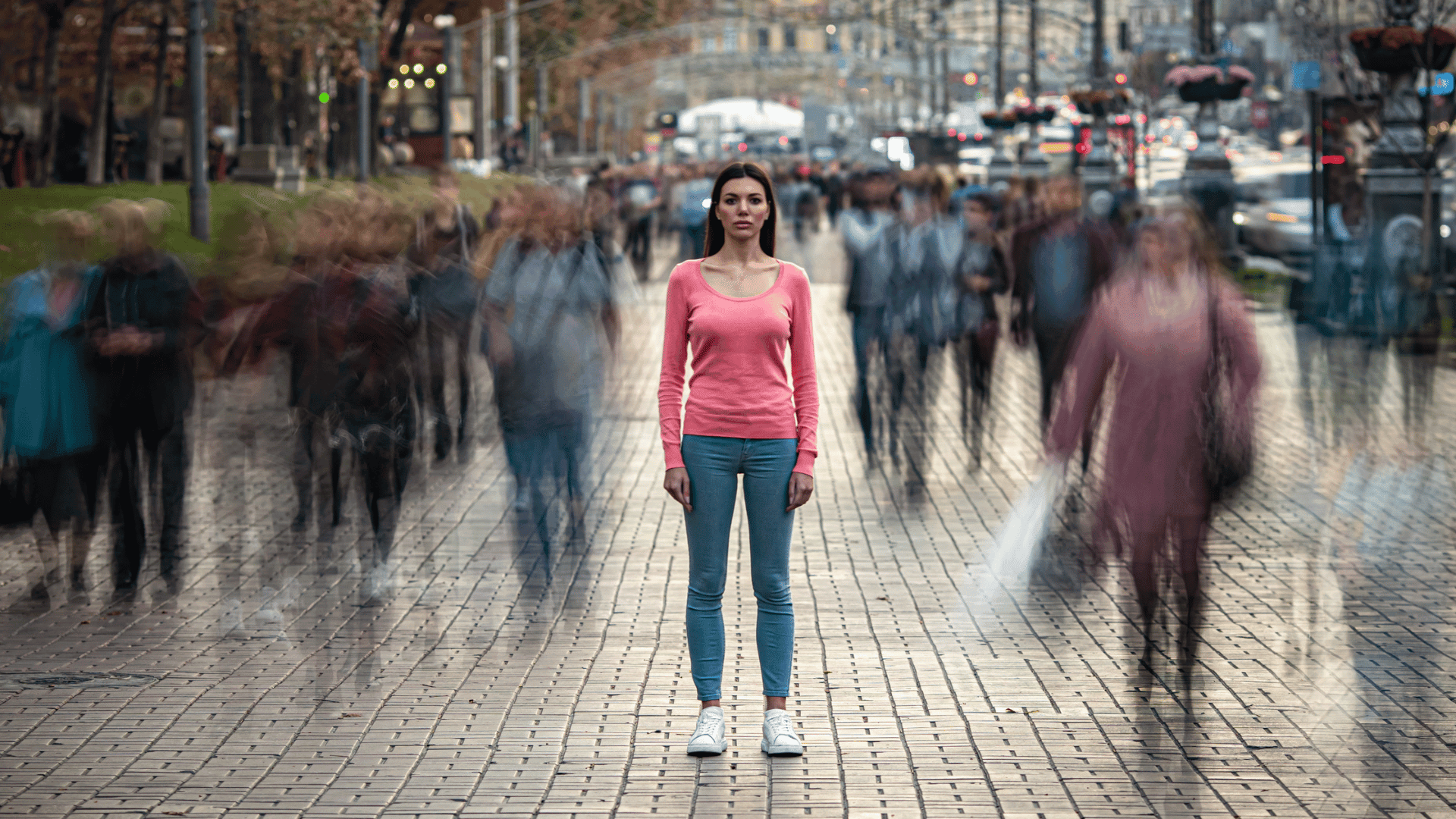 Woman in pink sweater standing still amidst a blurred crowd of moving people, representing the experience of feeling disconnected