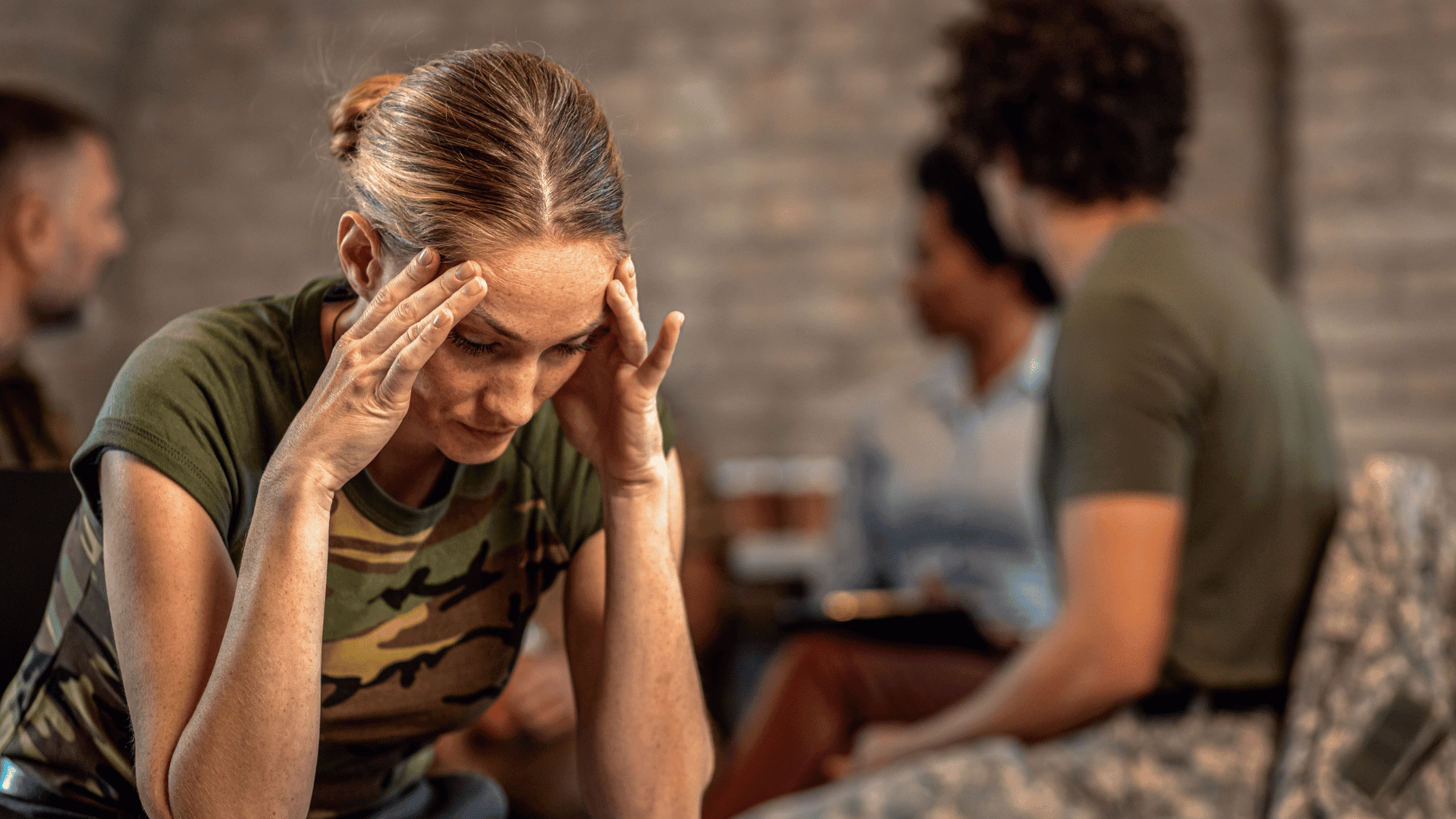 Woman in military camouflage shirt experiencing PTSD symptoms, holding her head while others are in the background